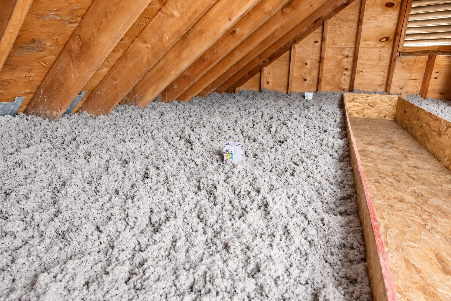 An attic floor is covered in thick gray blown-in insulation beneath exposed wooden rafters, with a narrow walkway along one side.