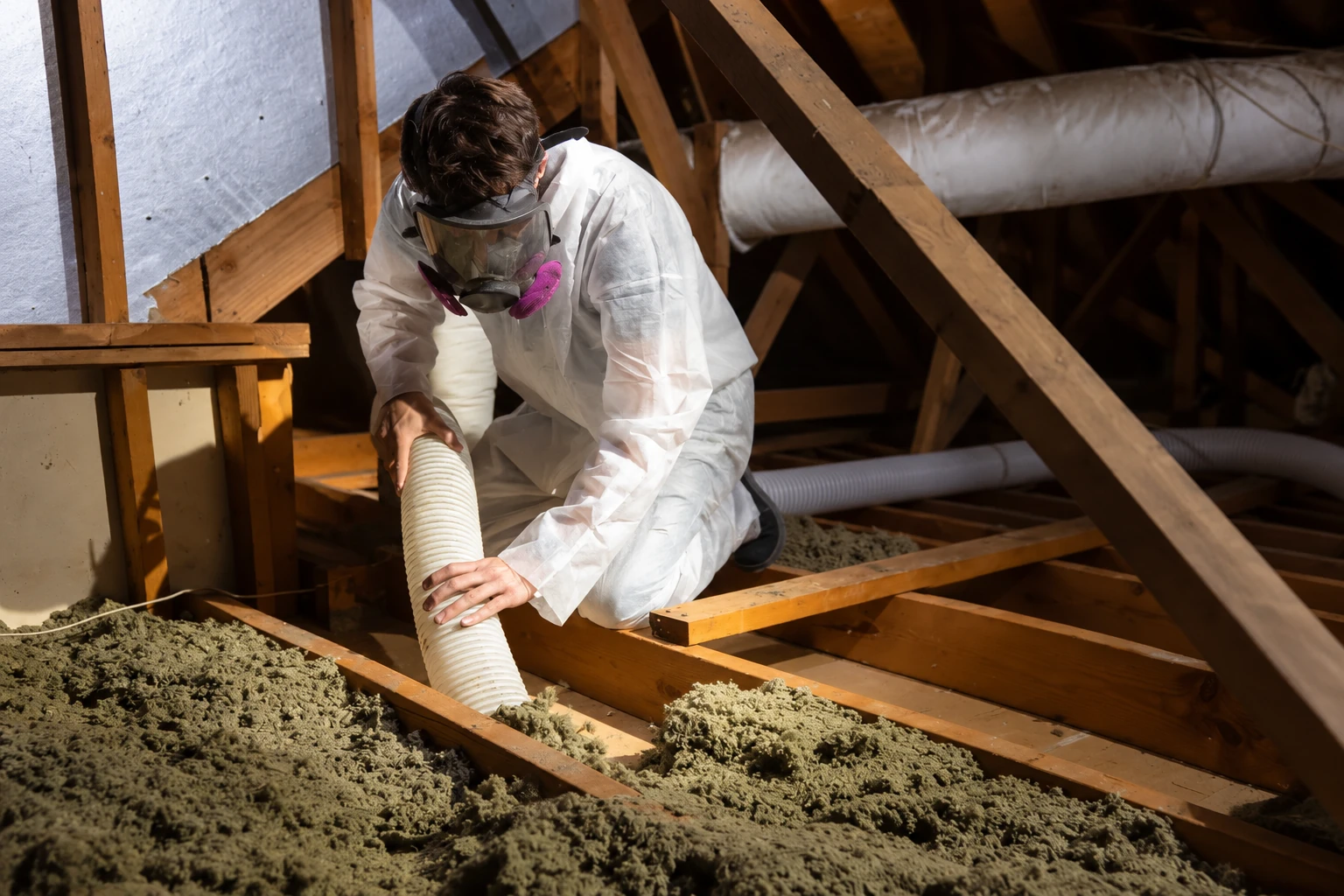 A worker in protective gear uses a hose to remove loose attic insulation between wooden joists.