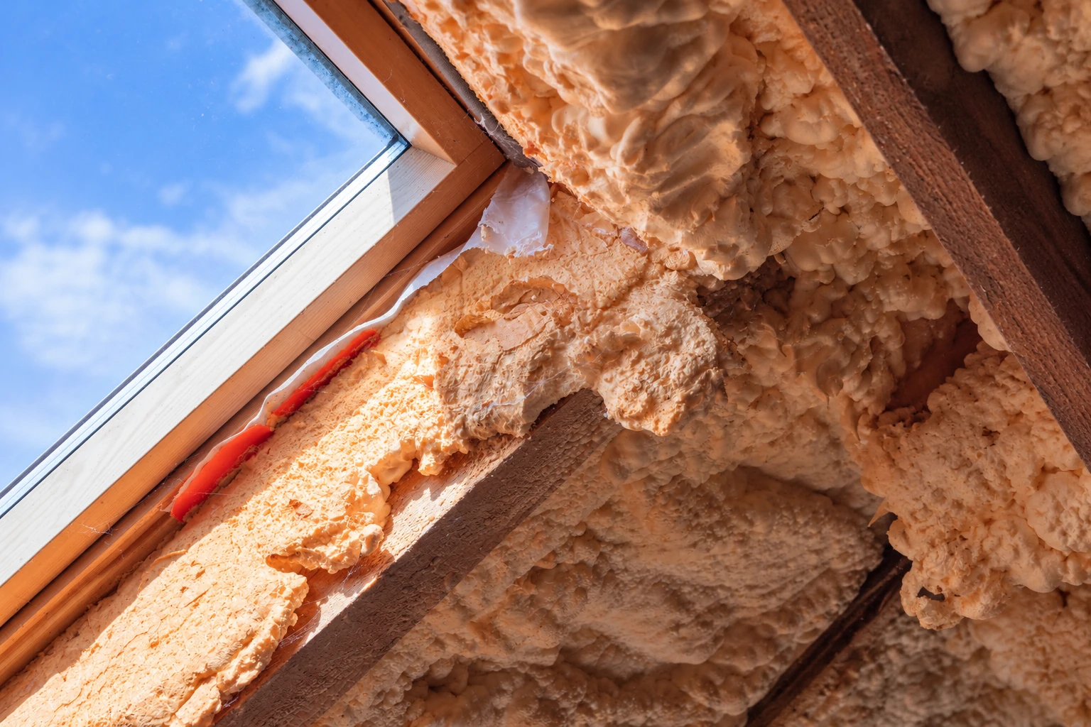 Open-cell spray foam fills the roof framing around a skylight, with blue sky visible through the glass.