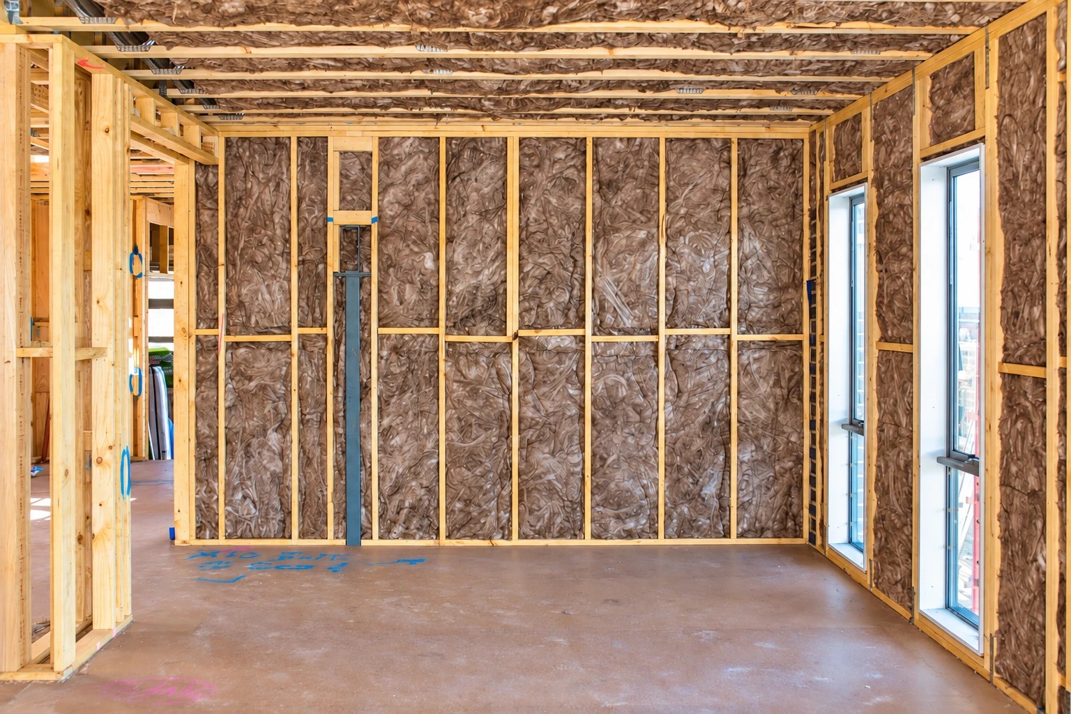 Fiberglass batts fill the exposed wall and ceiling framing of an unfinished room with tall windows.